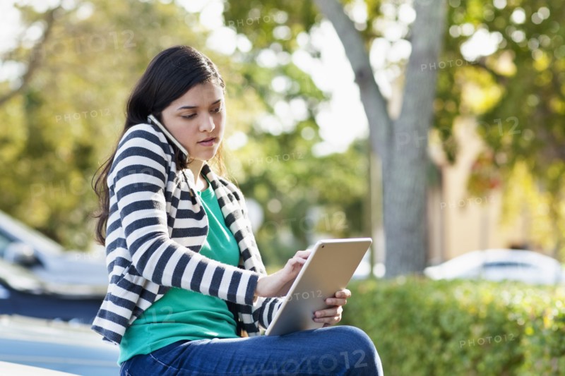 Hispanic woman using cell phone and digital tablet in parking lot