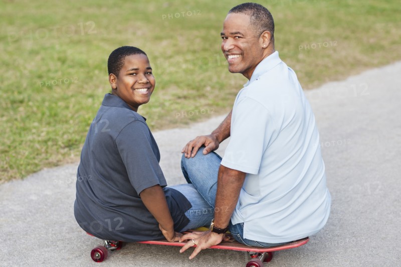 African American father and son sitting on skateboard on path