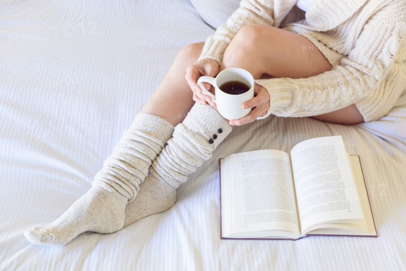 Caucasian woman reading book on bed and drinking coffee