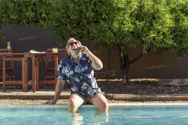 Caucasian man laughing on cell phone with legs in swimming pool