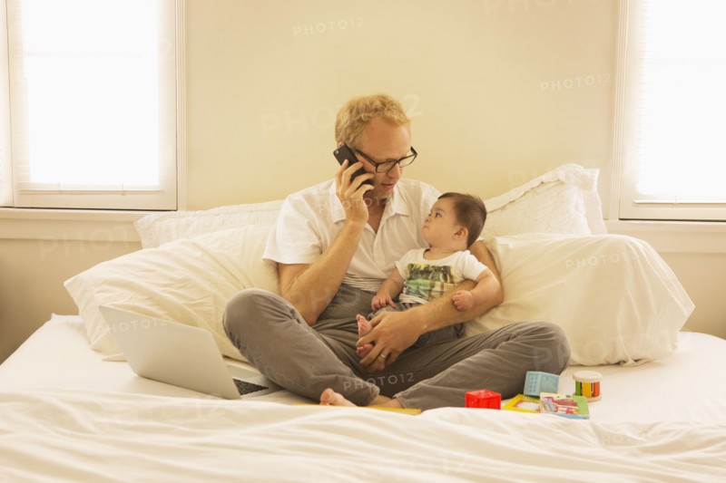 Father holding son and talking on cell phone on bed