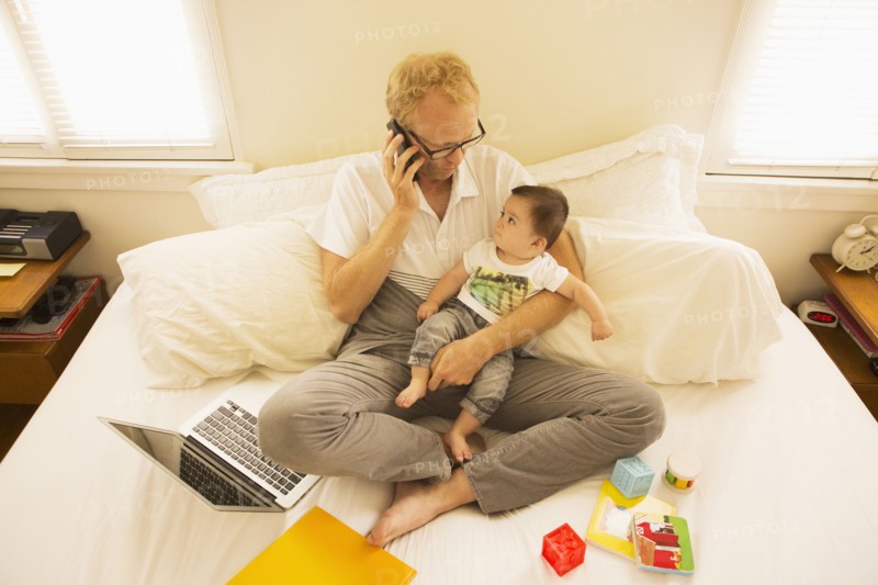 Father holding son and talking on cell phone on bed