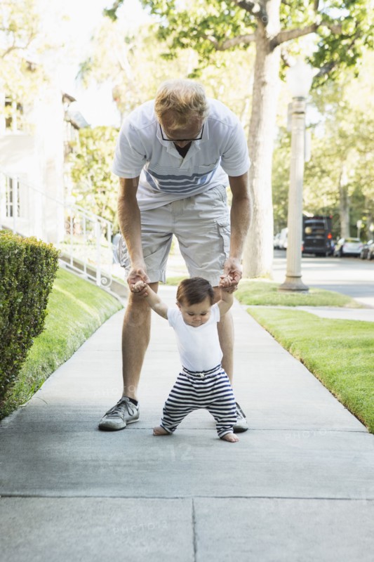 Father helping baby son walk on sidewalk