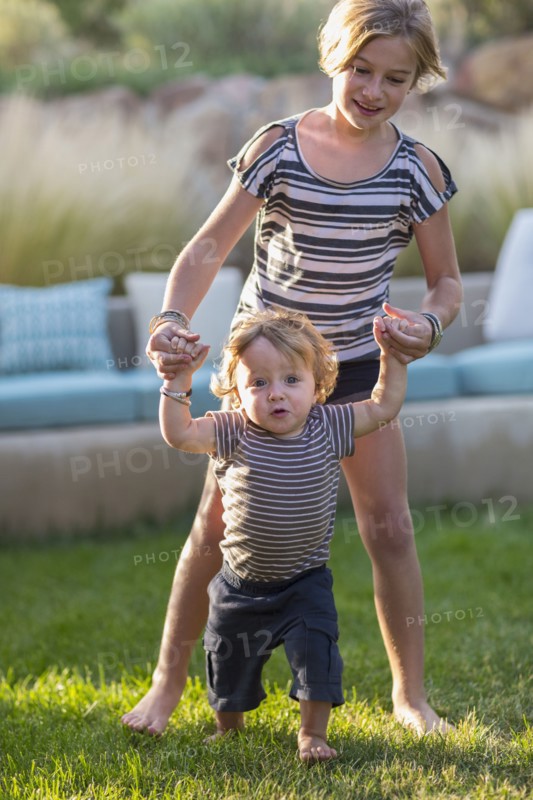 Caucasian sister helping baby brother walk in backyard