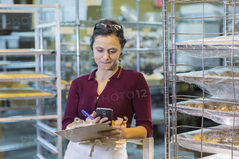Caucasian woman holding clipboard using cell phone in bakery