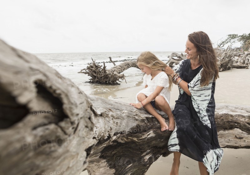 Caucasian mother and daughter sitting on driftwood on beach