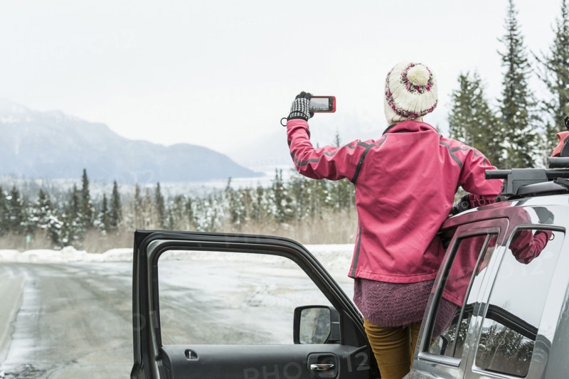 Caucasian woman standing in car in winter photographing scenic view
