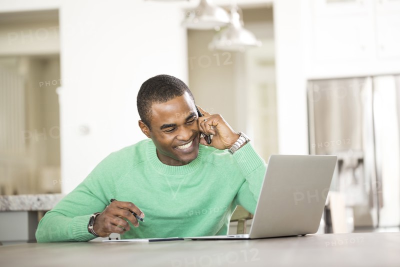 Black man sitting at table with laptop talking on cell phone