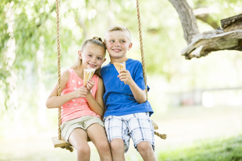 Caucasian brother and sister eating ice cream cones on rope swings