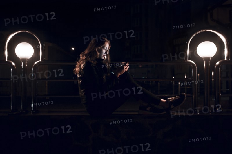 Caucasian teenage girl sitting on bench at night texting on cell phone