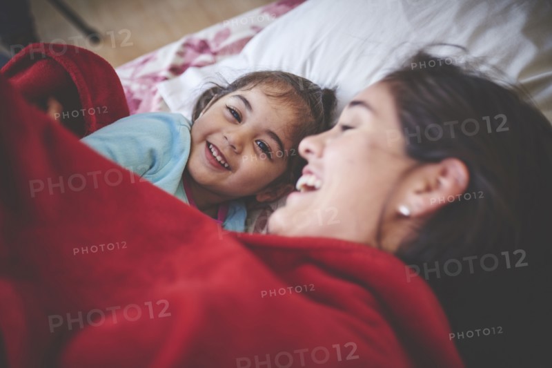 Laughing Hispanic mother and daughter under red blanket in bed