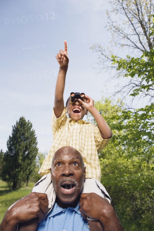 African grandfather carrying grandson on shoulders using binoculars in park