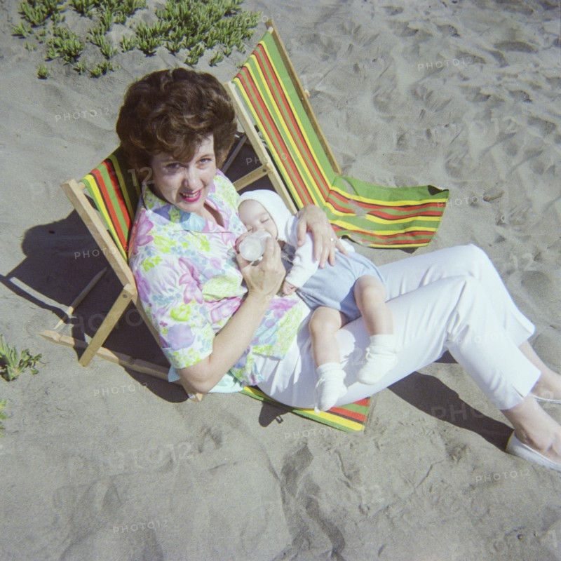 Caucasian mother sitting on beach feeding bottle to baby son
