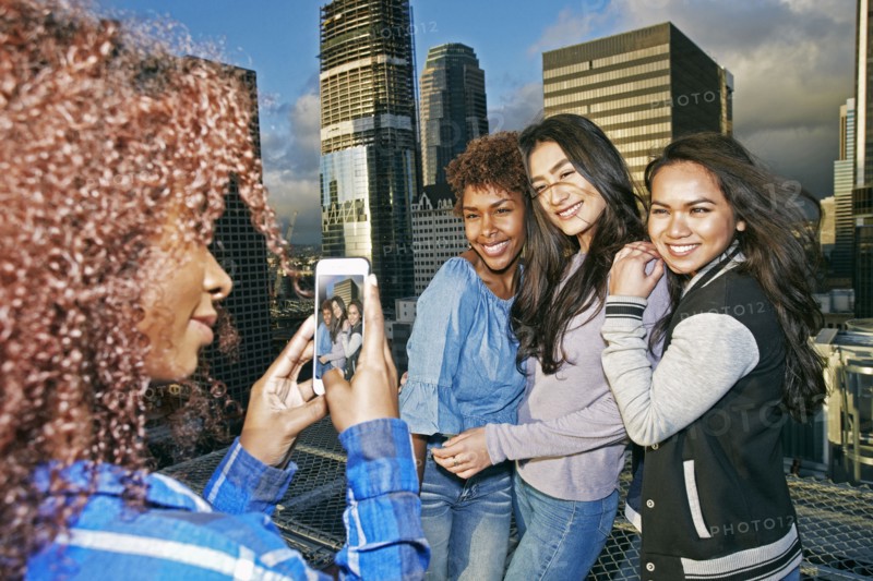 Stylish woman photographing friends on urban rooftop with cell phone