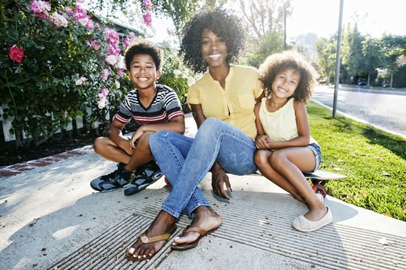 Smiling mother and children sitting on skateboard on sidewalk