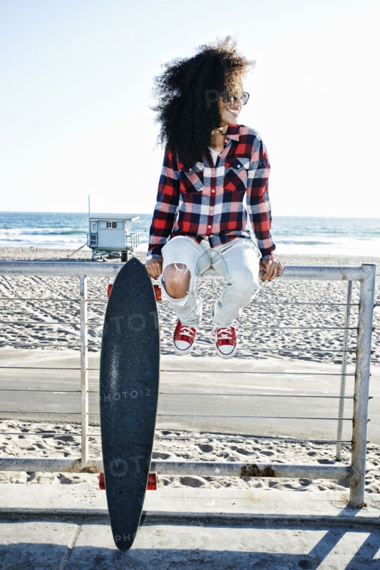 Hispanic woman sitting on fence at beach with skateboard