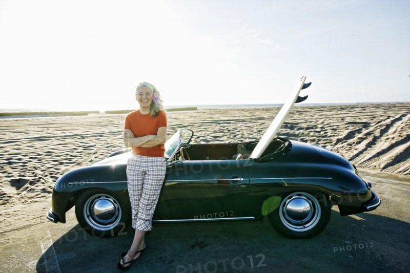 Older Caucasian woman leaning on convertible car with surfboard on beach