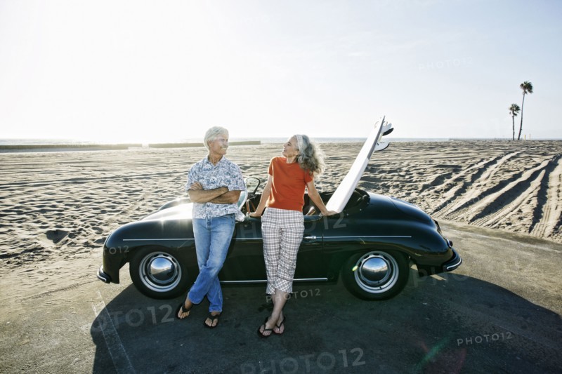 Older Caucasian couple leaning on convertible car with surfboard on beach