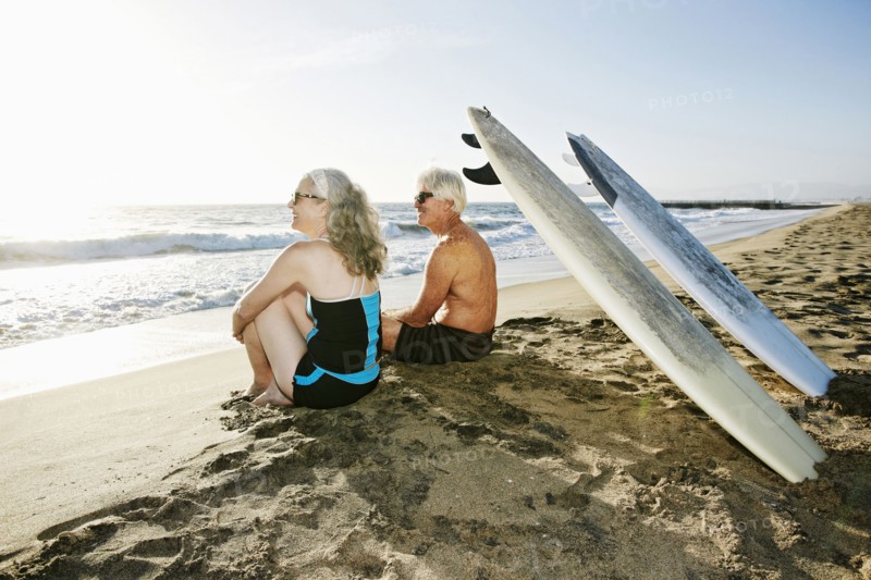 Older Caucasian couple sitting on beach with surfboards