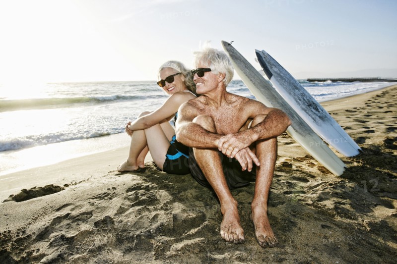 Older Caucasian couple sitting on beach with surfboards
