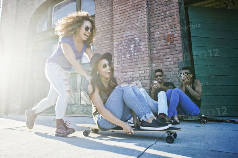 Woman pushing friend sitting on skateboard in city