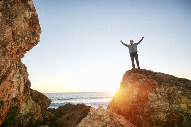 Caucasian man standing with arms raised on rock at ocean