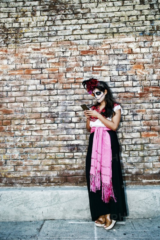 Mixed Race woman on sidewalk wearing skull face paint texting on cell phone