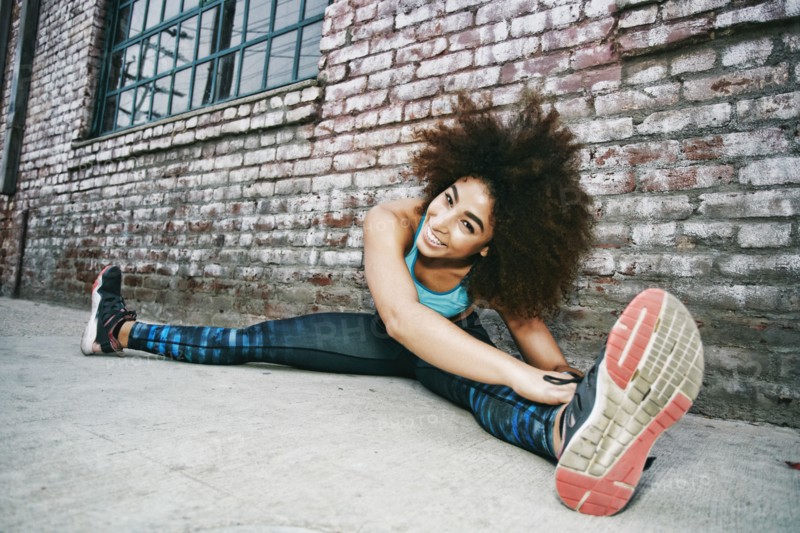 Hispanic woman sitting on sidewalk stretching legs near brick wall