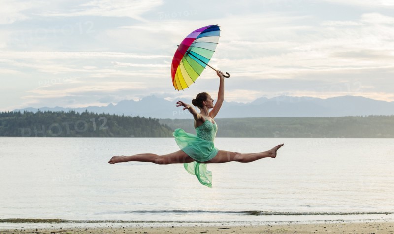 Caucasian ballerina jumping with multicolor umbrella on beach