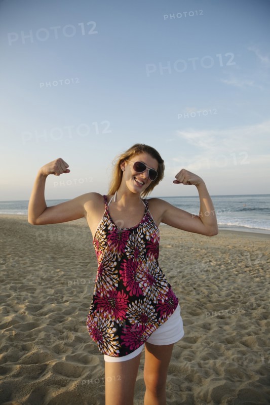 Smiling Caucasian teenage girl flexing biceps on beach