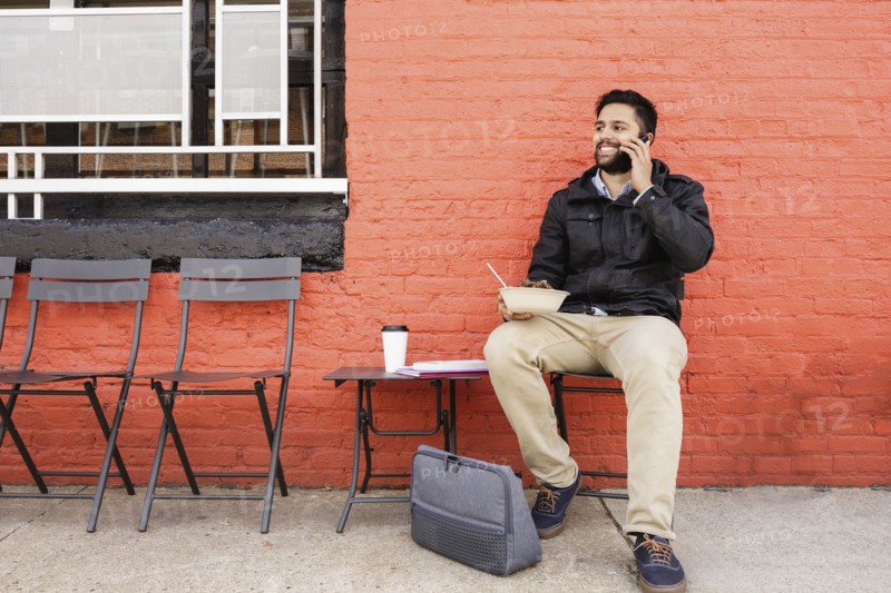 Hispanic man eating bowl of food on sidewalk using cell phone