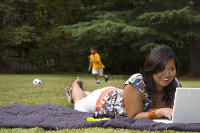Asian mother using laptop in park with son in background