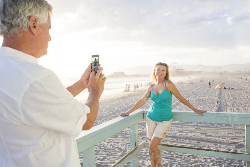 Caucasian man taking photograph of wife on boardwalk at beach