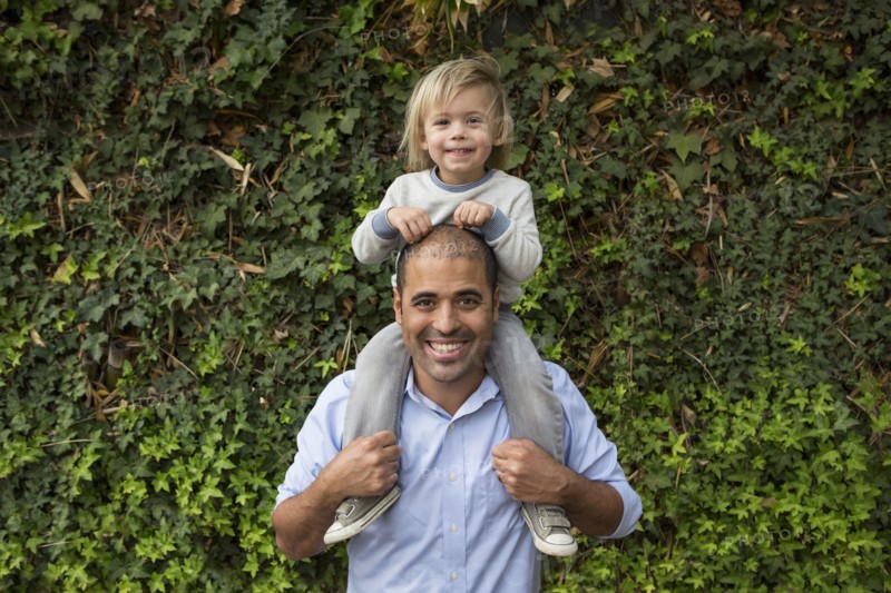Hispanic father carrying son on shoulders near ivy wall