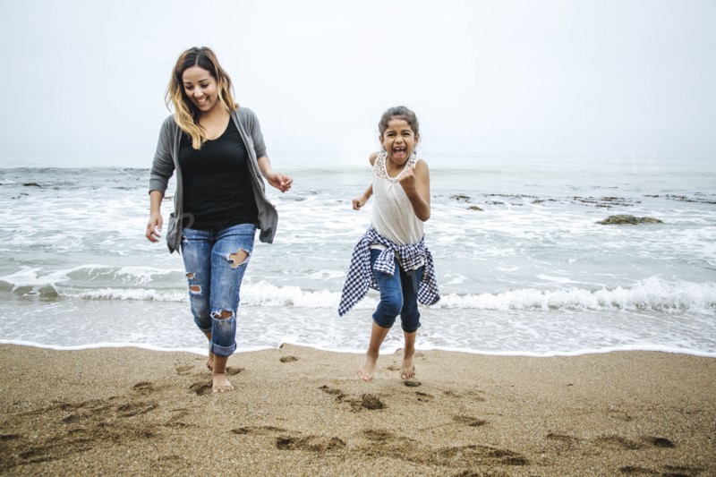 Hispanic mother and daughter walking barefoot on beach