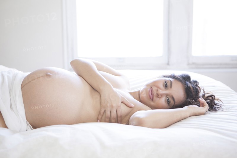 Nude Hispanic pregnant woman covering her breasts on bed