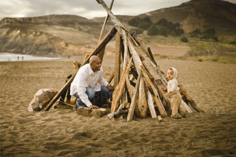 Father and baby son sitting under driftwood teepee on beach