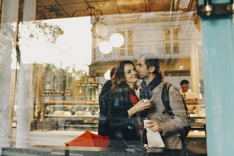 Caucasian man kissing woman on cheek behind bakery window