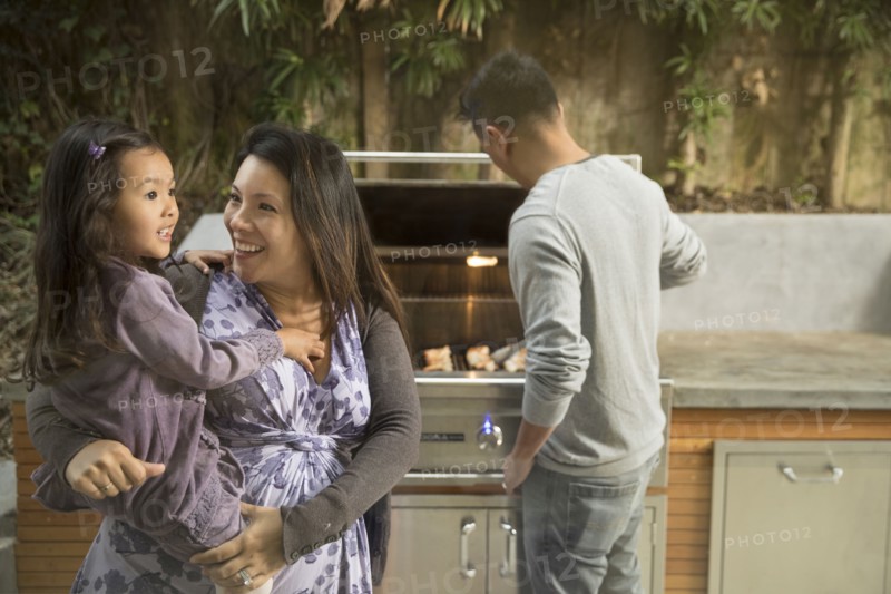 Expectant mother carrying daughter while husband is cooking on grille