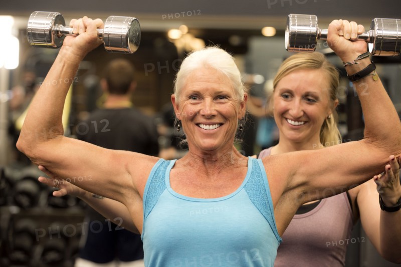 Trainer assisting older woman lifting dumbbells in gymnasium