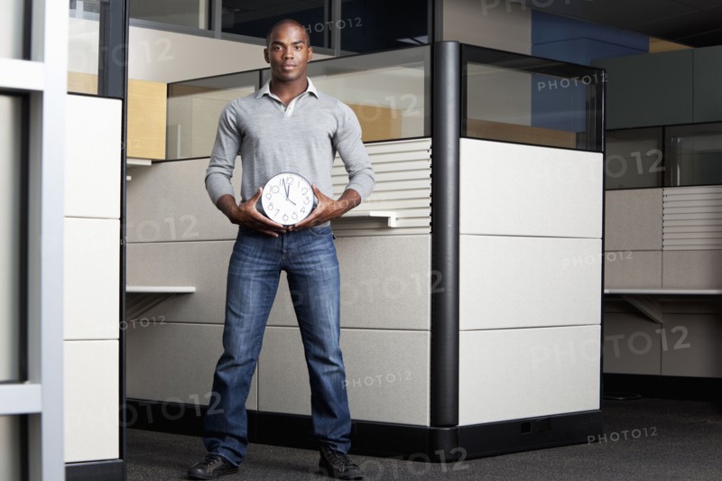 Mixed race businessman holding clock in office cubicle