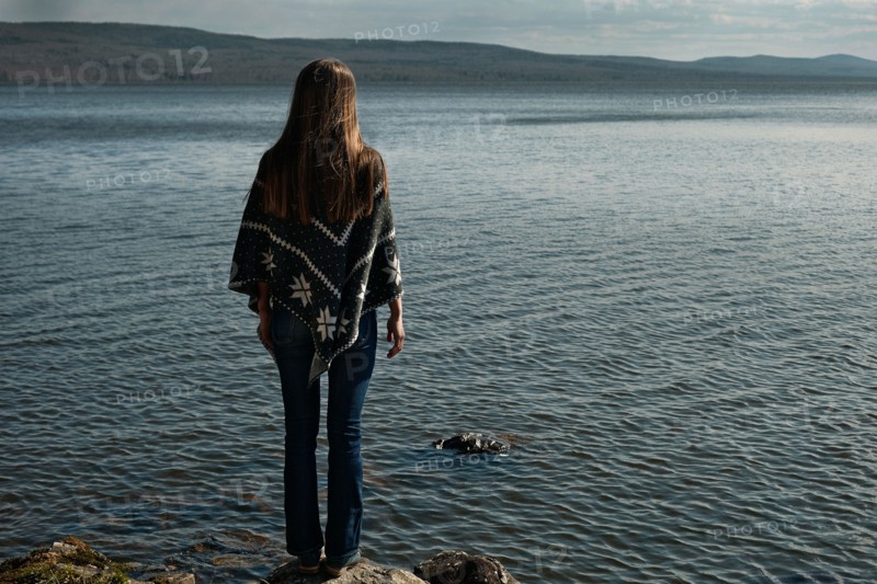 Pensive Caucasian girl standing on rock at lake