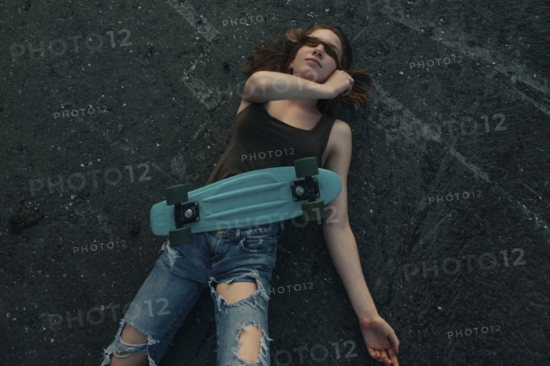 Caucasian teenage girl with skateboard laying on pavement covering eyes