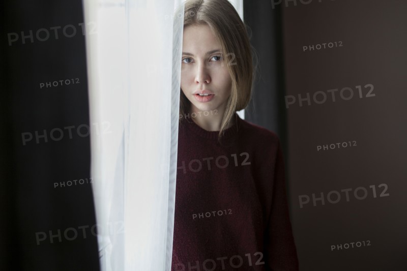 Serious Caucasian woman standing near curtain at window
