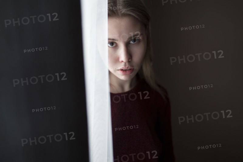 Serious Caucasian woman standing near curtain at window