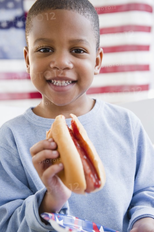 African American boy eating hot dog in front of American flag