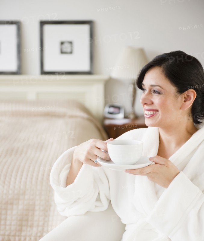 Hispanic woman in bathrobe drinking coffee in bedroom