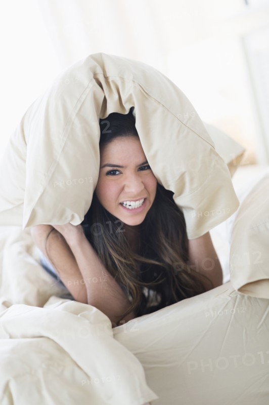 Portrait of frustrated Hispanic woman under pillow in bed