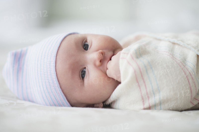 Swaddled Asian baby biting fingers on bed