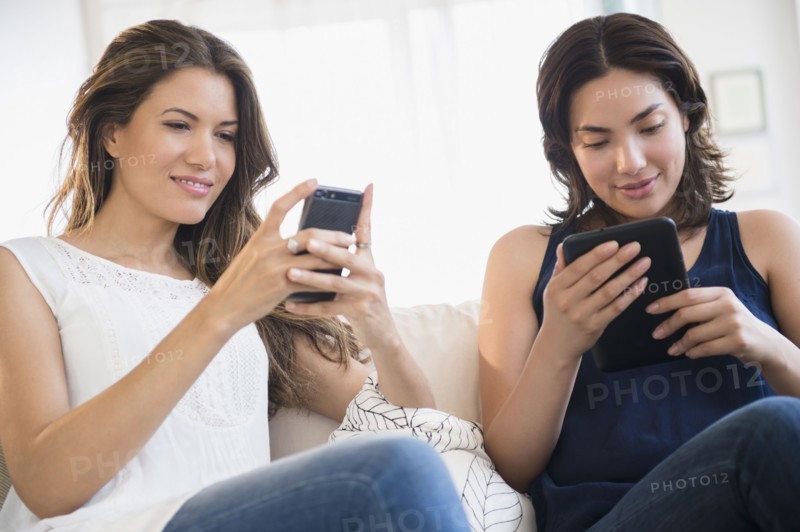 Hispanic women using cell phone and digital tablet on sofa